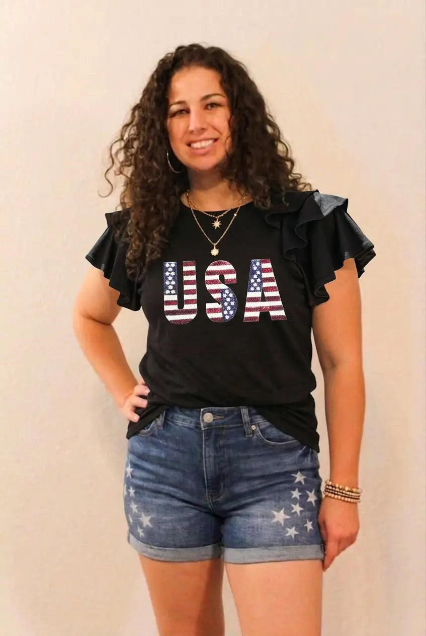 Woman wearing a black 'USA' t-shirt with ruffled sleeves and denim shorts with star patterns on a plain background.